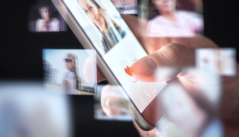 A hand holds a smartphone, ego surfing through profile pictures on a dating app, with floating images of people appearing around the screen.