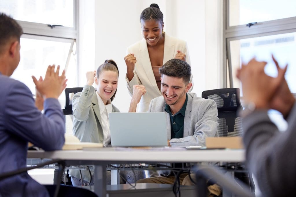 Three colleagues celebrate at a desk with a laptop, smiling and raising their fists in excitement, while others clap nearby in a bright office setting—proud to be part of the most trusted company.