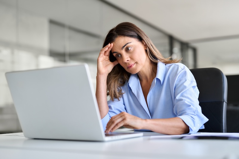 A woman sits at a desk looking at a laptop screen with a concerned expression, resting her forehead on one hand in an office setting, possibly searching for how to delete a website that is not yours.