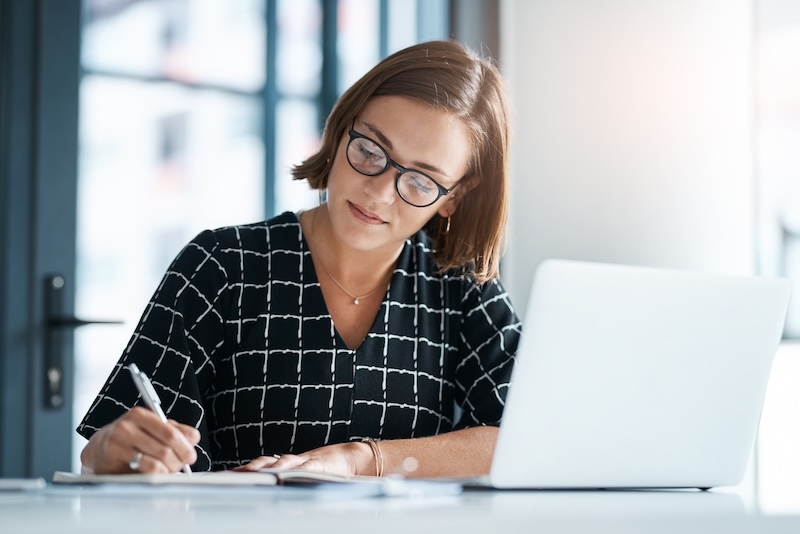 A woman wearing glasses and a patterned top is jotting down notes in her notebook at a desk. Seated in front of an open laptop, with sunlight streaming through the window, she wonders silently, "Can I make a Wikipedia page for myself?.