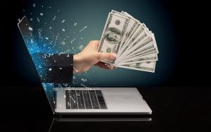 A hand holding a laptop with money flying out of it.