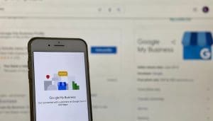 Smartphone displaying the "Google My Business" app screen, featuring a logo with colorful business icons. In the blurred background, a Google My Business webpage is open on a computer screen.