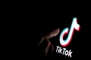 A hand is reaching toward a glowing TikTok logo on a dark background, highlighting the app's signature red, blue, and white musical note icon above the text "TikTok"—evoking questions about reputation risk in the digital age.