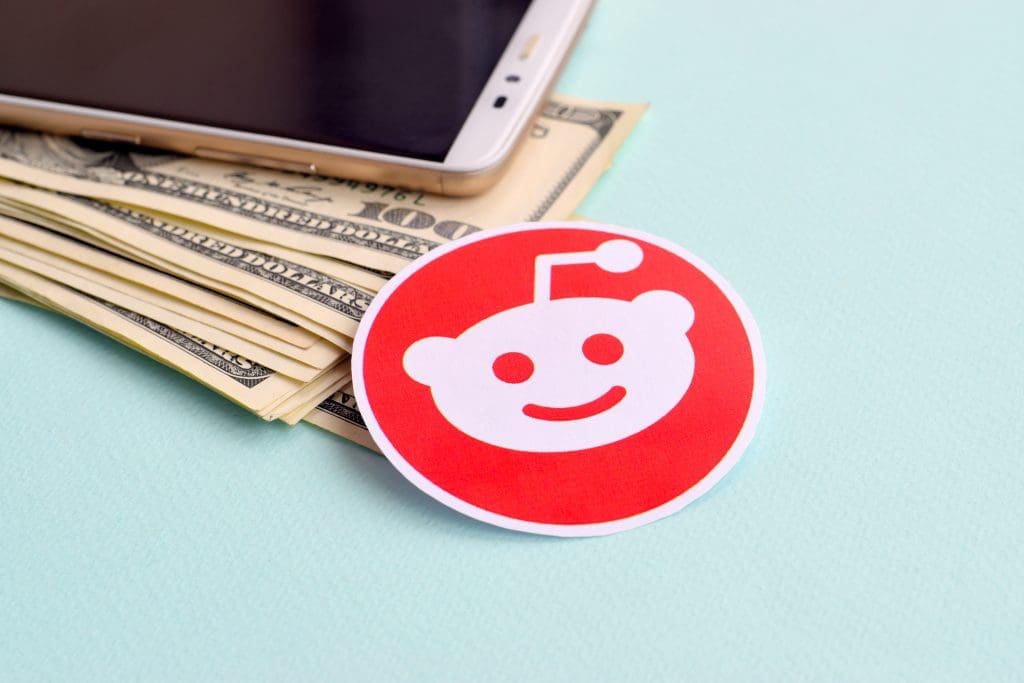 A stack of US dollar bills with a smartphone placed on top, next to a round sticker featuring the red and white Reddit logo, all on a light blue surface.