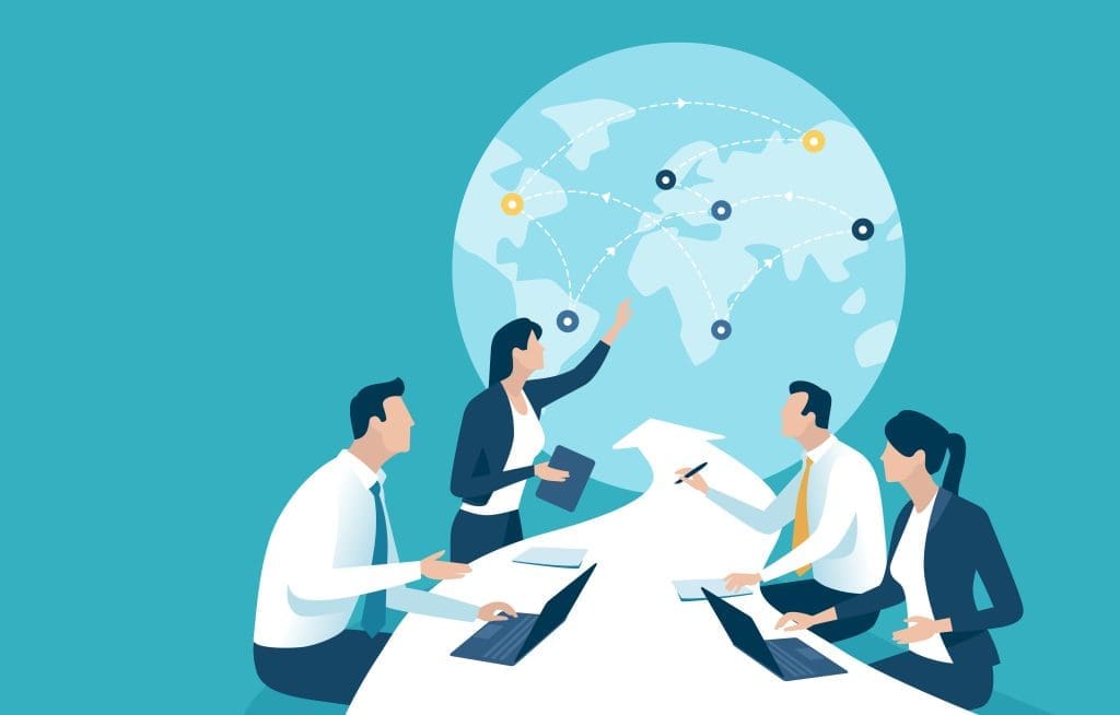 Four business professionals sit at a conference table with laptops, discussing, while a woman points to a world map with connected locations in the background, representing global teamwork or strategy.