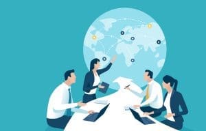 Four business professionals sit at a conference table with laptops, discussing, while a woman points to a world map with connected locations in the background, representing global teamwork or strategy.