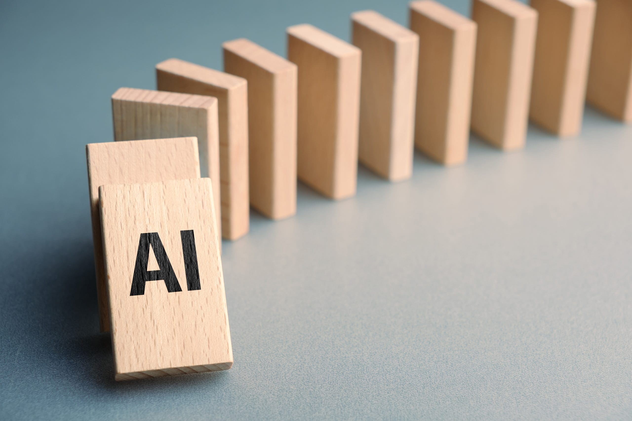 A row of standing wooden dominoes, with the first one in the foreground labeled "AI." The dominoes are positioned in a way that suggests a chain reaction.