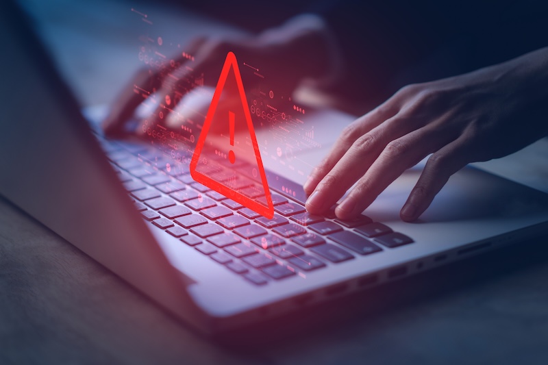 Hands typing on a laptop keyboard with a large red warning triangle and exclamation mark symbol digitally superimposed, highlighting an online safety alert or computer security warning.
