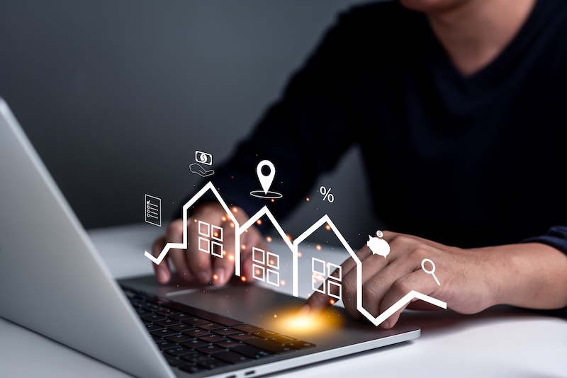 A person types on a laptop with digital icons of houses, a location pin, percentage, magnifying glass, and piggy bank floating above the keyboard, highlighting how realtors need online reputation management for property investment or search.