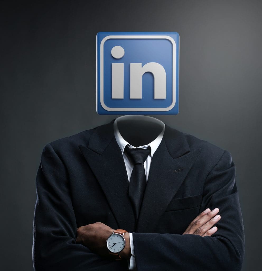 A person in a suit with arms crossed, but instead of a head, there is a large LinkedIn logo. The background is dark and plain.