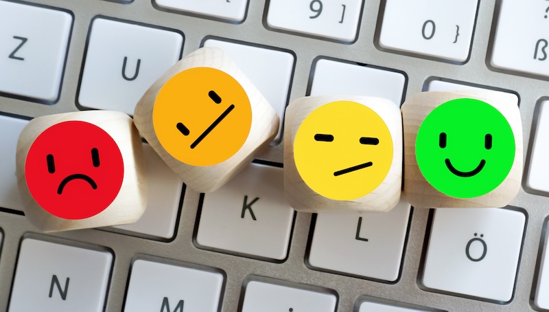 Four wooden blocks with colorful emoji faces—red sad, orange neutral, yellow indifferent, and green happy—are placed in a row on a computer keyboard, representing emotions like those analyzed when Google detects fake reviews.