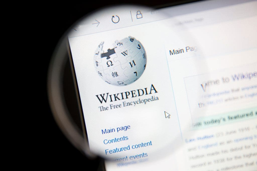A magnifying glass hovers over a computer screen displaying the Wikipedia logo, text "Wikipedia The Free Encyclopedia," and a menu with links like Main page, Contents, and Featured content.