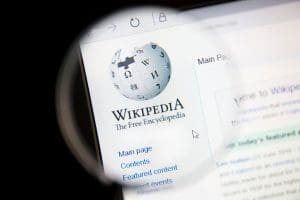 A magnifying glass hovers over a computer screen displaying the Wikipedia logo, text "Wikipedia The Free Encyclopedia," and a menu with links like Main page, Contents, and Featured content.