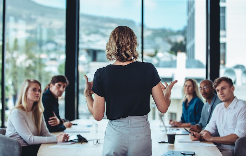 A woman stands and speaks to five colleagues seated around a conference table in a modern office with large windows and a city view, leading a discussion on total corporate social responsibility as the group listens attentively during the meeting.