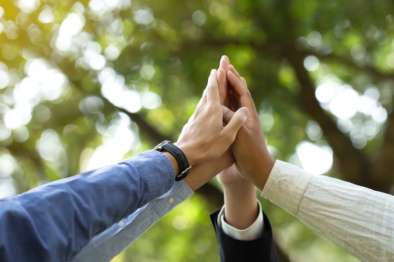 Four hands coming together for a group high-five outdoors, with blurred green trees and sunlight in the background, symbolizing teamwork and total corporate social responsibility.