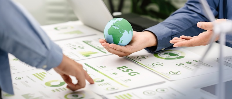 Two people in business attire discuss ESG documents on a desk, with one person holding a small globe, highlighting total corporate social responsibility and a commitment to sustainability.