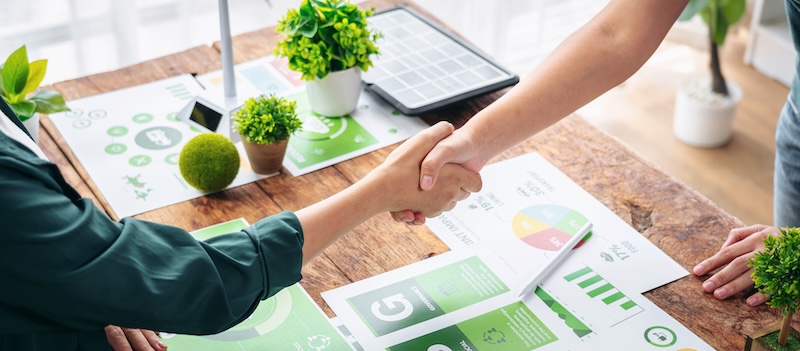 Two people shake hands over a desk covered with green-themed charts, graphs, plants, and a solar panel, suggesting a meeting focused on sustainability or total corporate social responsibility initiatives.