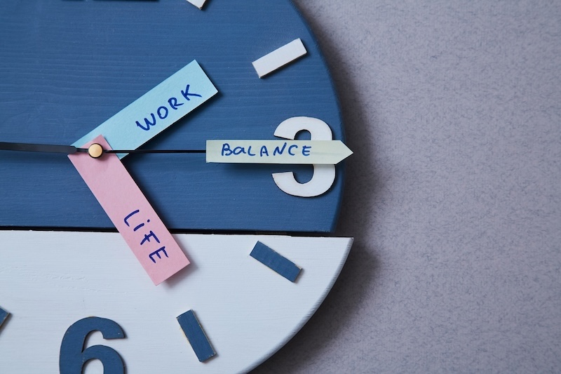 A clock with paper labels as clock hands: "WORK" and "LIFE" point at different times, and "BALANCE" stretches across the face, symbolizing work-life balance—a visual nod to total corporate social responsibility.