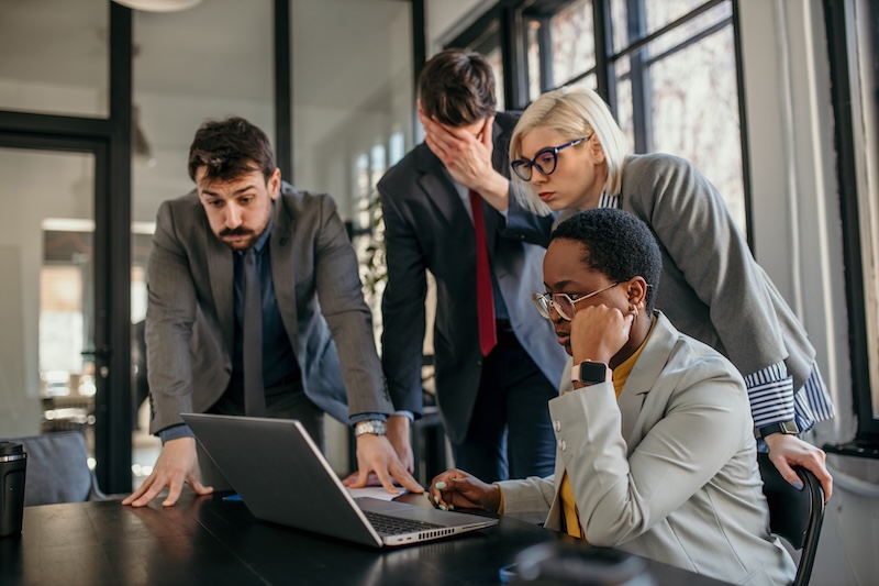 Four business professionals gather around a laptop, looking stressed and concerned after receiving negative news. One person sits in front of the laptop with a hand on their face, while the others stand closely behind, observing the screen.