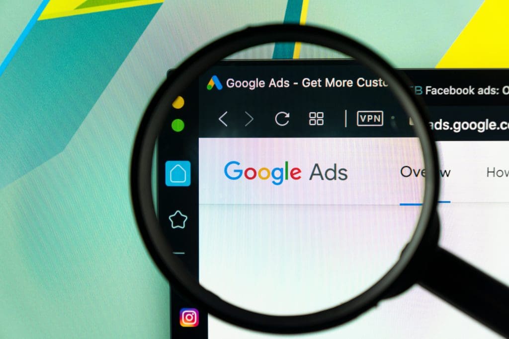 A computer screen displays the Google Ads website, with the words “Google Ads” clearly visible. A magnifying glass is held over the screen, emphasizing the Google Ads logo and part of the browser interface.