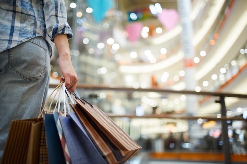 Close-up of a person standing in a mall, holding bags and looking at stores in the distance.