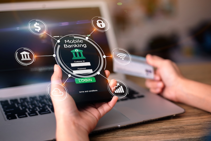 A person uses a laptop and a smartphone with a mobile banking app. They hold a credit card in their other hand. Different mobile banking icons show around the phone.
